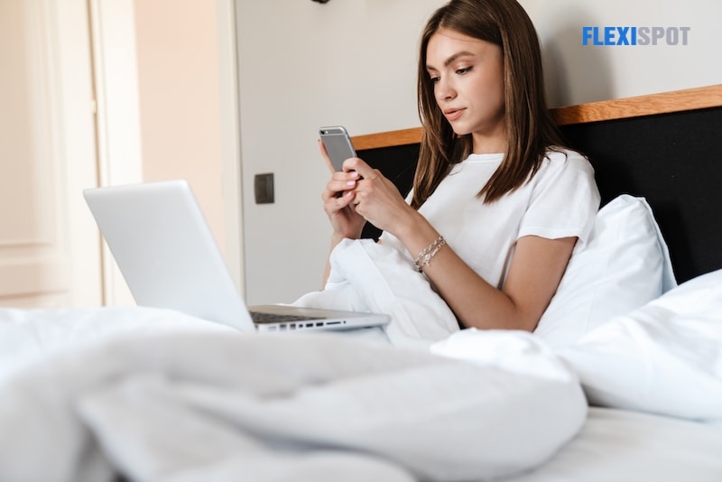 Attractive young woman using mobile phone and laptop computer while relaxing in bed