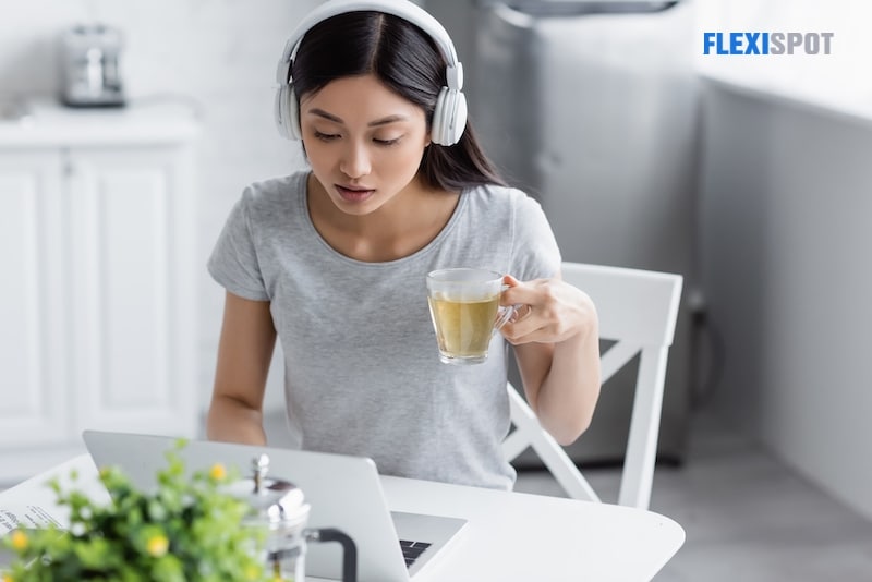 Young asian woman in headphones looking at laptop while holding cup of tea
