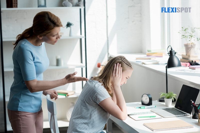 Depressed teenage daughter sitting at the desk at home while her mother yelling at her