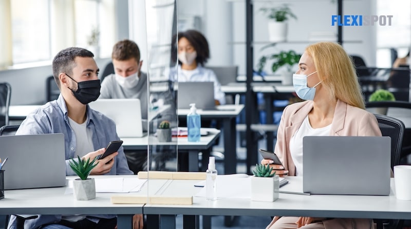 Young men and women wearing protective masks, holding smartphones, holding laptops and preservatives in the workplace, talking through protective glass.