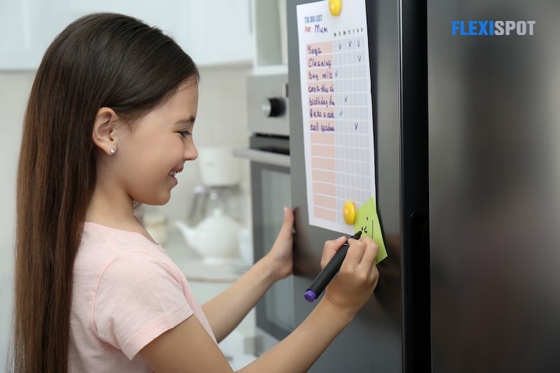 Little girl drawing funny face on note near to do list in kitchen