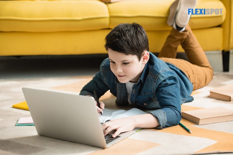 Attentive boy lying on the floor next to the book, using laptop while doing homework