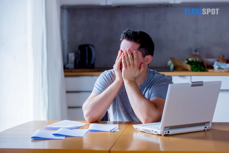 Young tired man holding his head in hand looking for a job online, sitting in the kitchen holding a laptop.